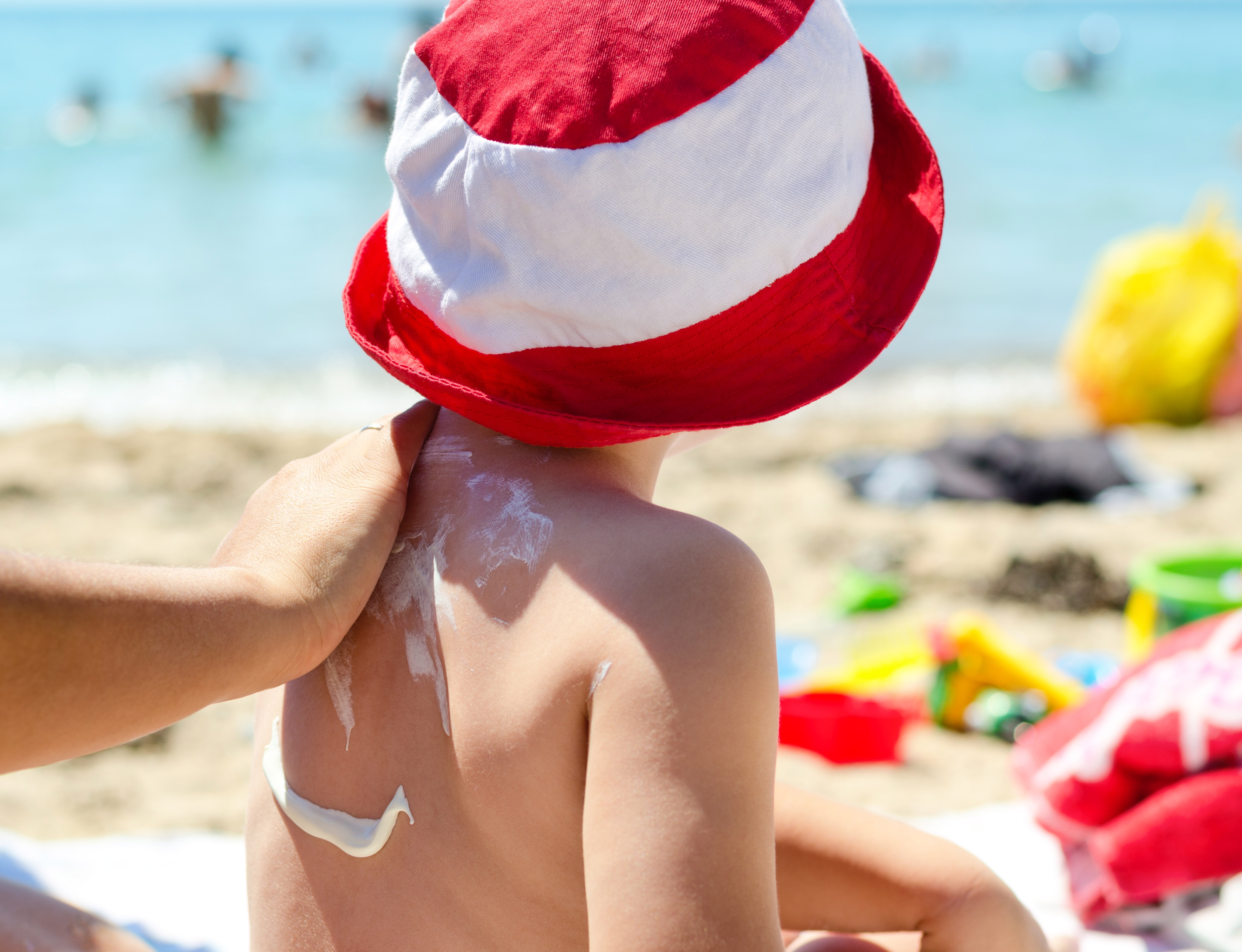 Young boy sitting on the beach during his summer vacation having sunscreen applied to his back by a parent to prevent burning from harmful UV rays