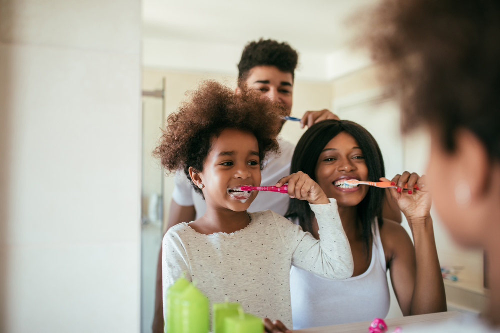 Mother, father, and child brush teeth in the mirror.