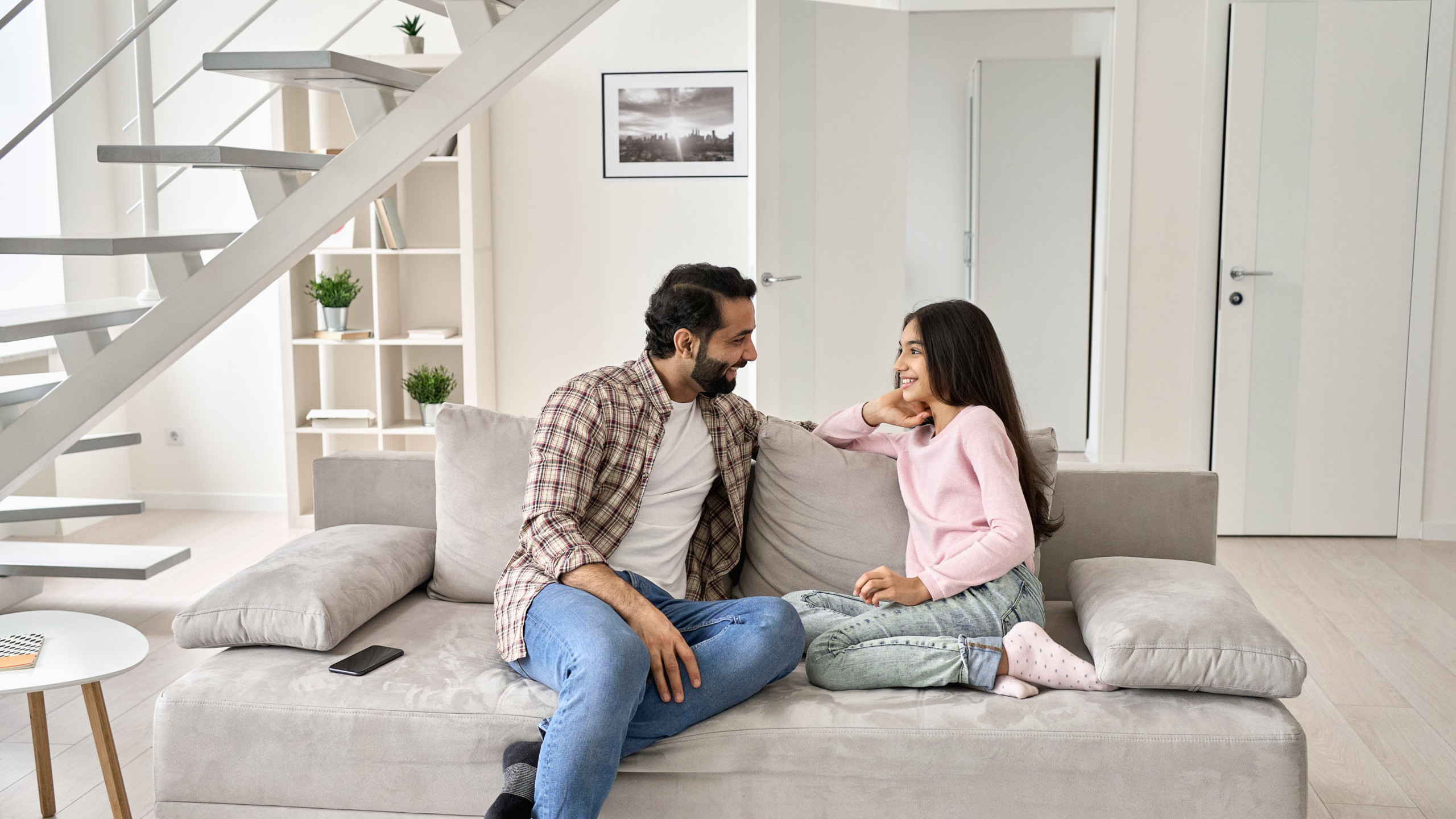 Happy young indian father with teenage child daughter having fun enjoying talking spending time at home. Smiling dad and teen kid girl having conversation sitting on sofa in modern house living room.
