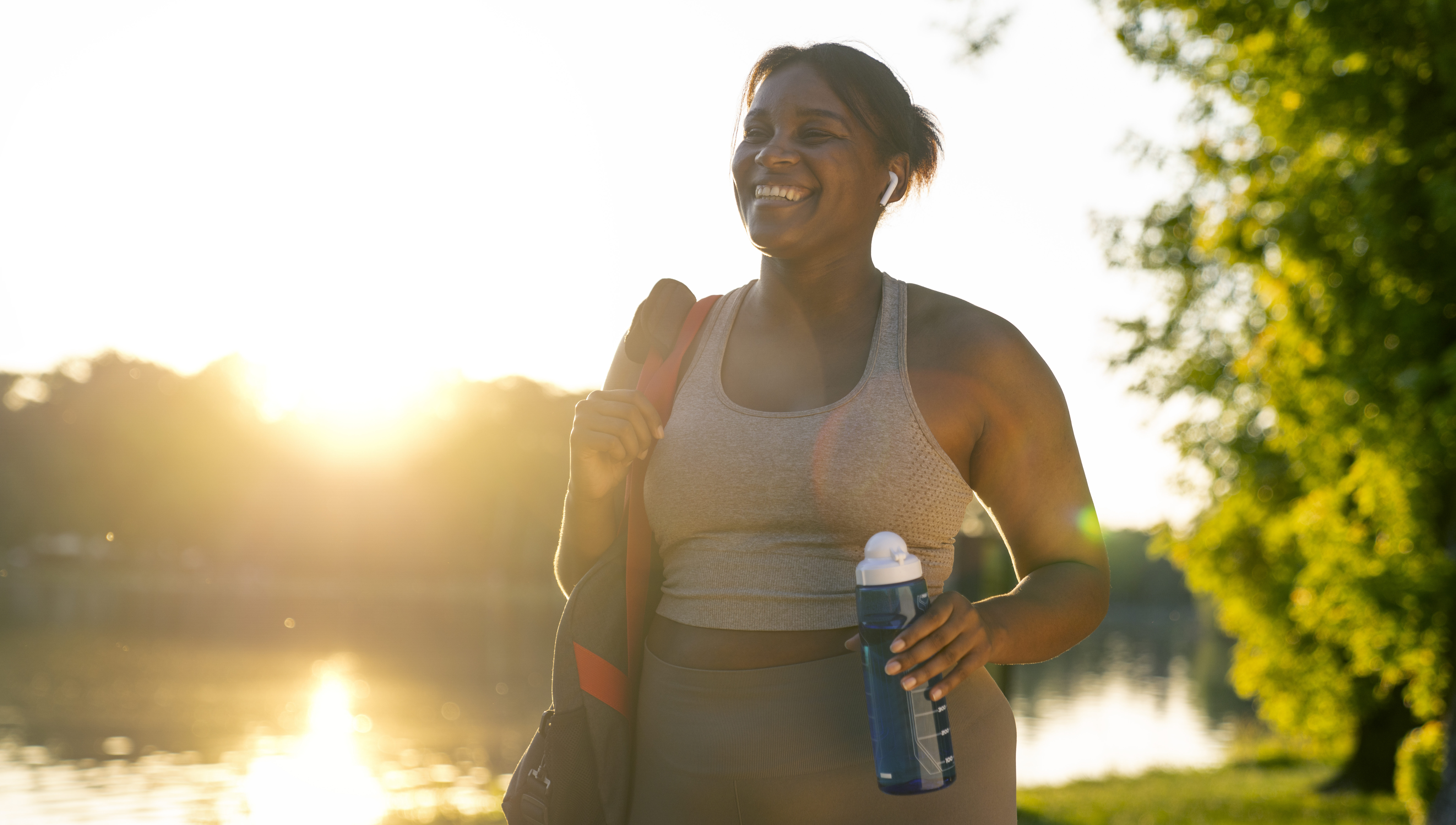 Young African American woman walking with bag and bottle of water through the park in a summer day