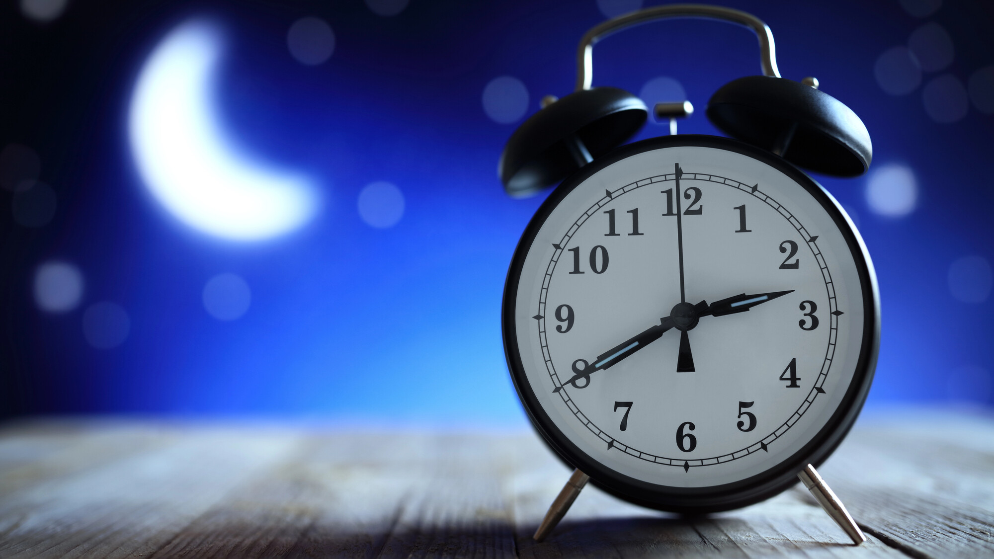 Alarm analog desk clock sits on a wood desk in front of a mural depicting a crescent moon and starts over a dark blue night sky