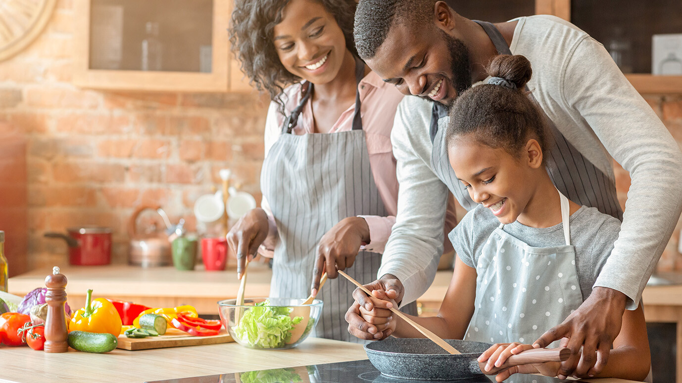 Black parents guide their daughter in cooking healthy food in a sunlit kitchen.