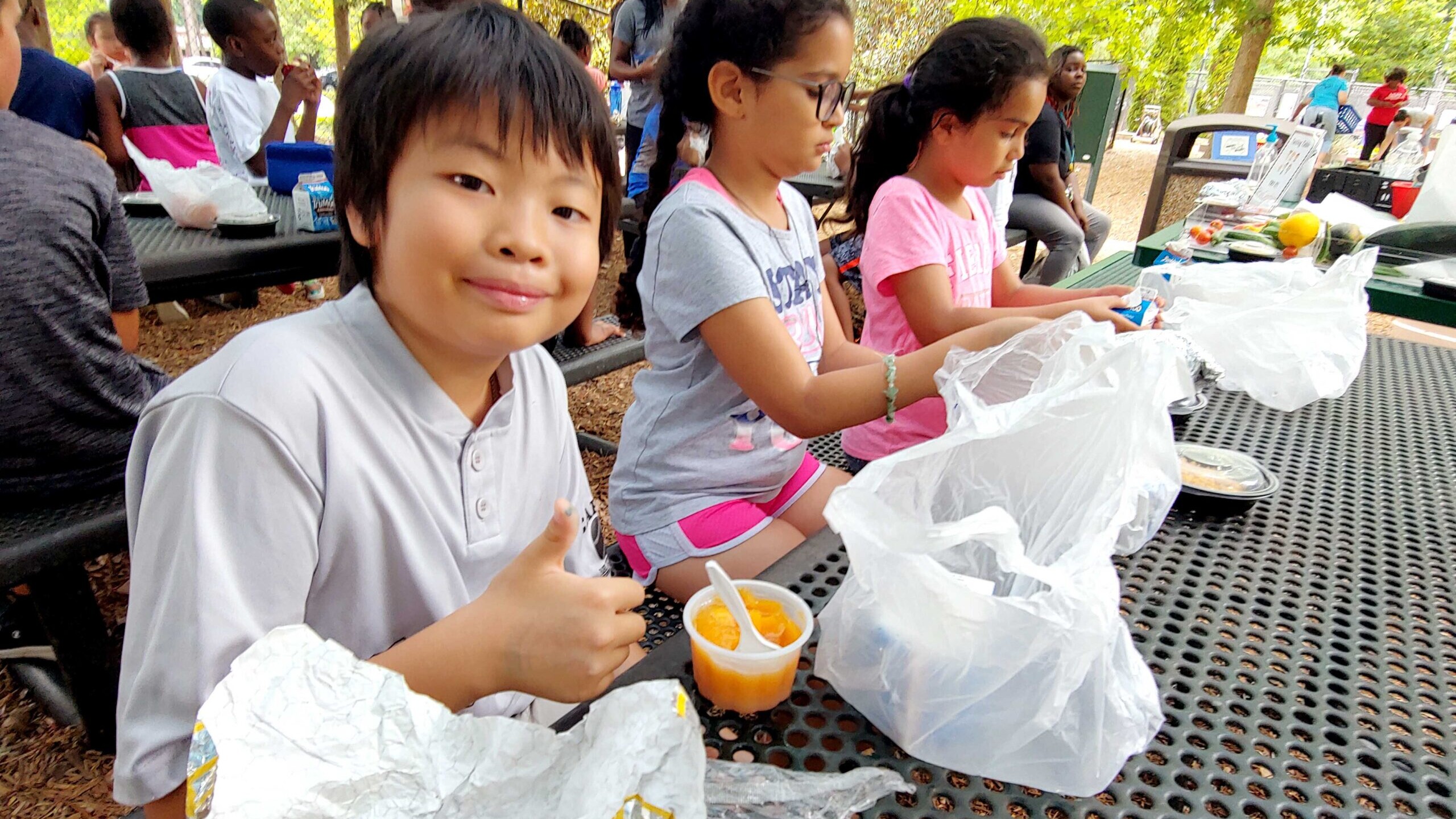 Young participants of the Summer Meals program at the Poe Center smile at the camera as they eat nutritious lunches outside on a picnic table.