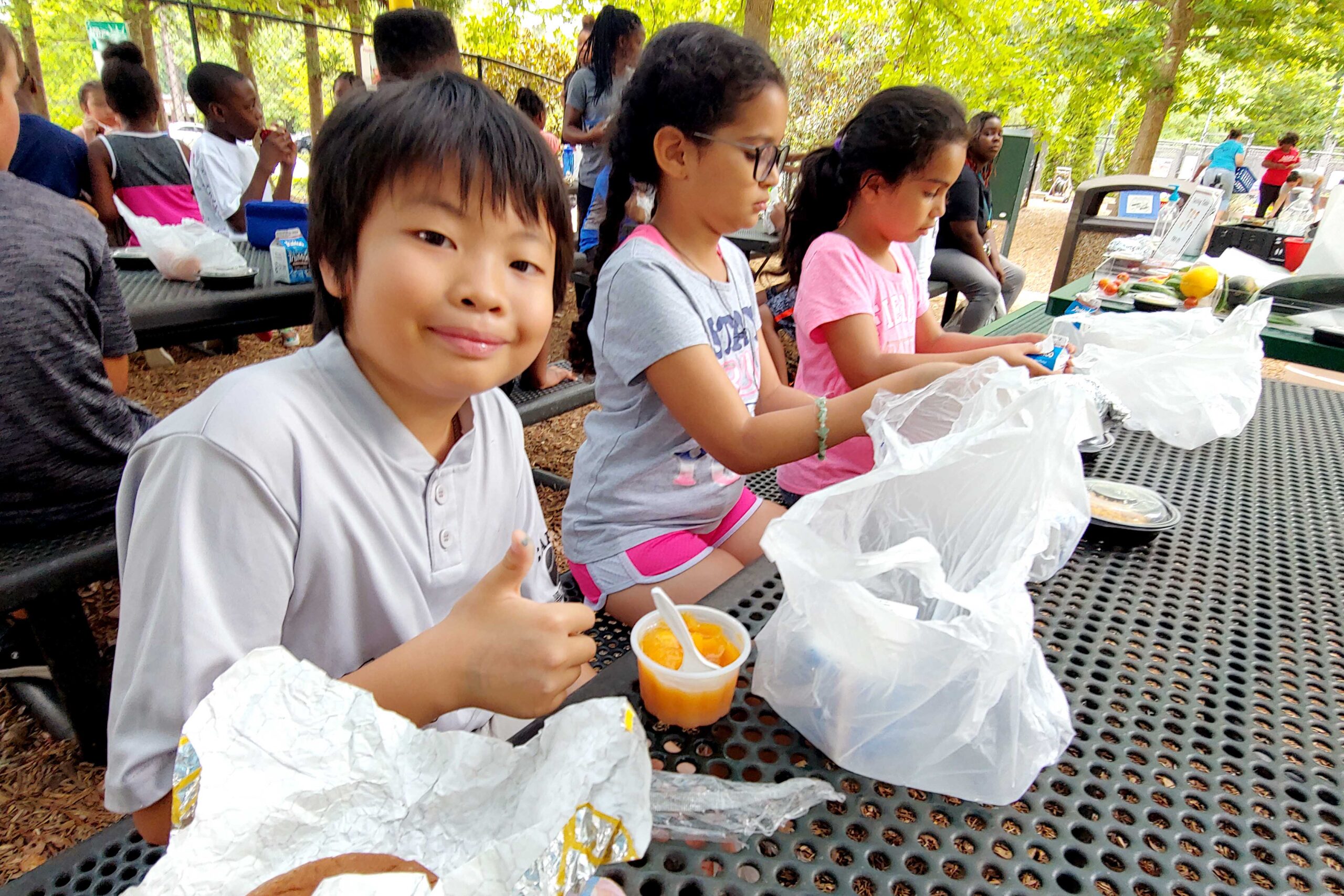 Young participants of the Summer Meals program at the Poe Center smile at the camera as they eat nutritious lunches outside on a picnic table.