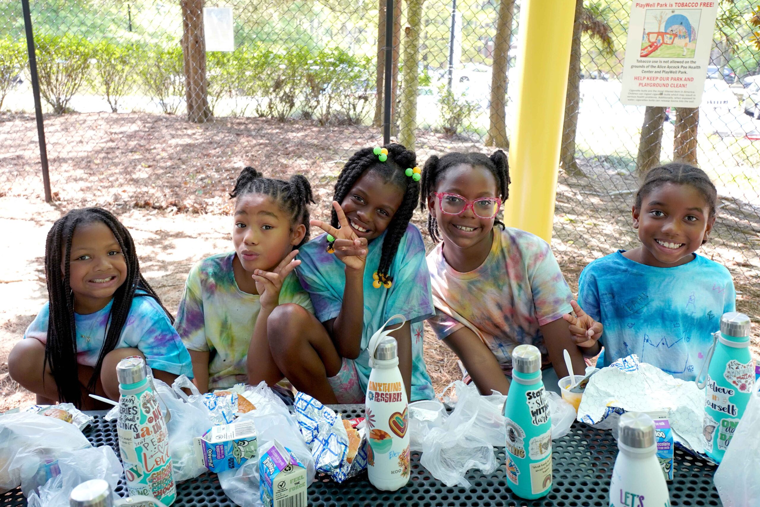 Five young girl participants of Summer Meals at the Poe Center smile at the camera as they eat nutritious lunches outside on a picnic table.