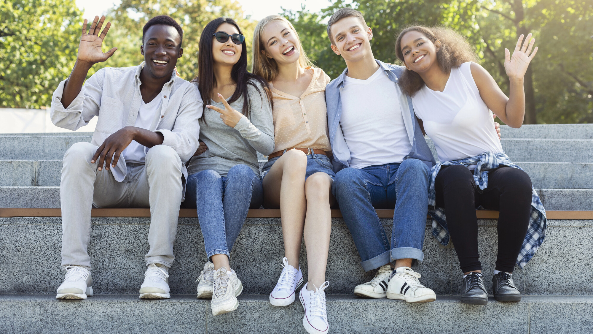 five diverse friends sit on stone steps outside and wav at the camera
