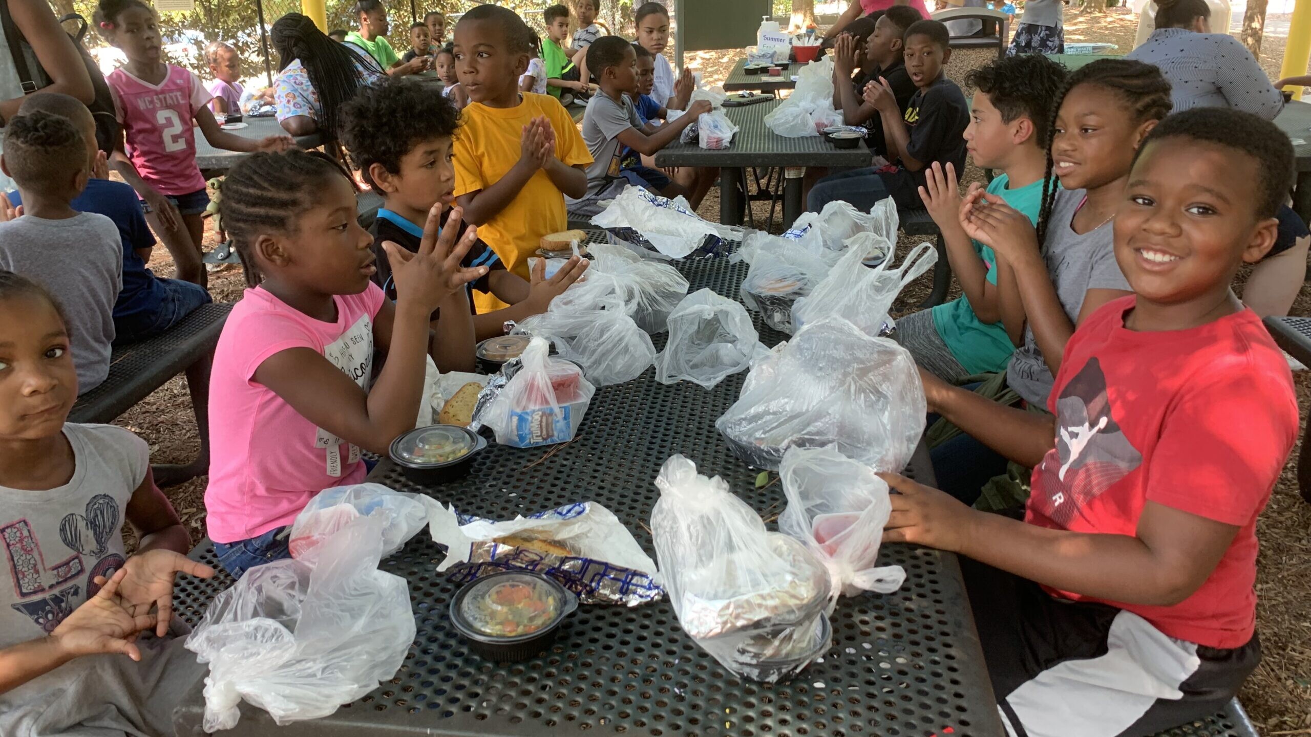 Several children sit at a gray picnic table and smile at the camera during a Summer Meals event at the Poe Center