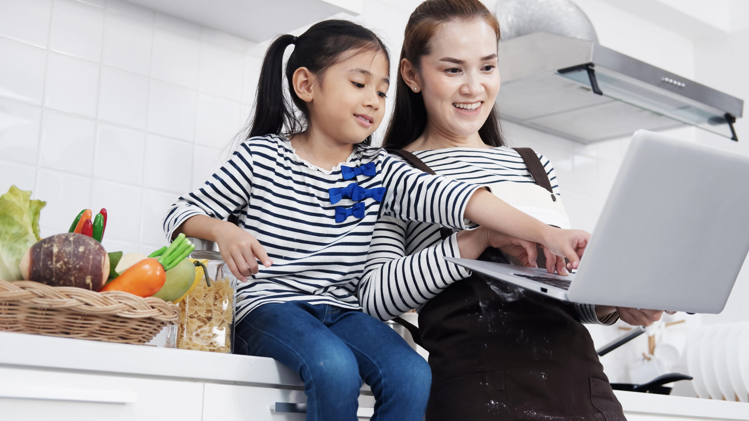 mother and daughter sit at kitchen counter and look at a laptop screen