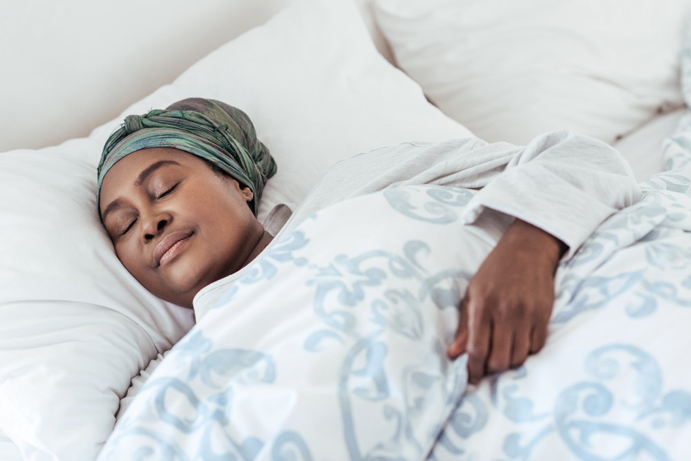 A middle aged woman wearing a silk bonnet lies in bed sleeping under a blue comforter