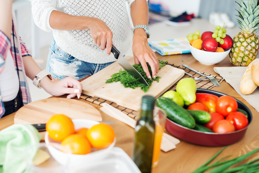 Two women stand at a kitchen table preparing a healthy meal full of vegetables.